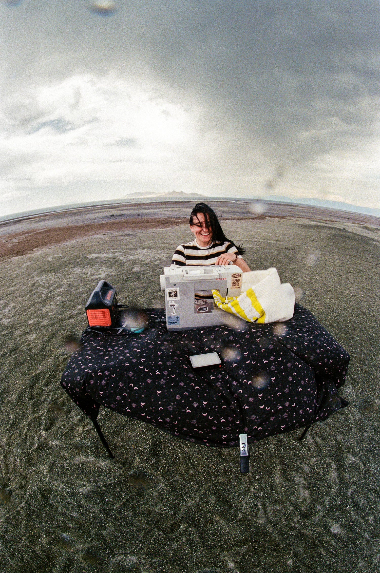 Person sitting on a round platform with a sewing machine in a desert-like setting